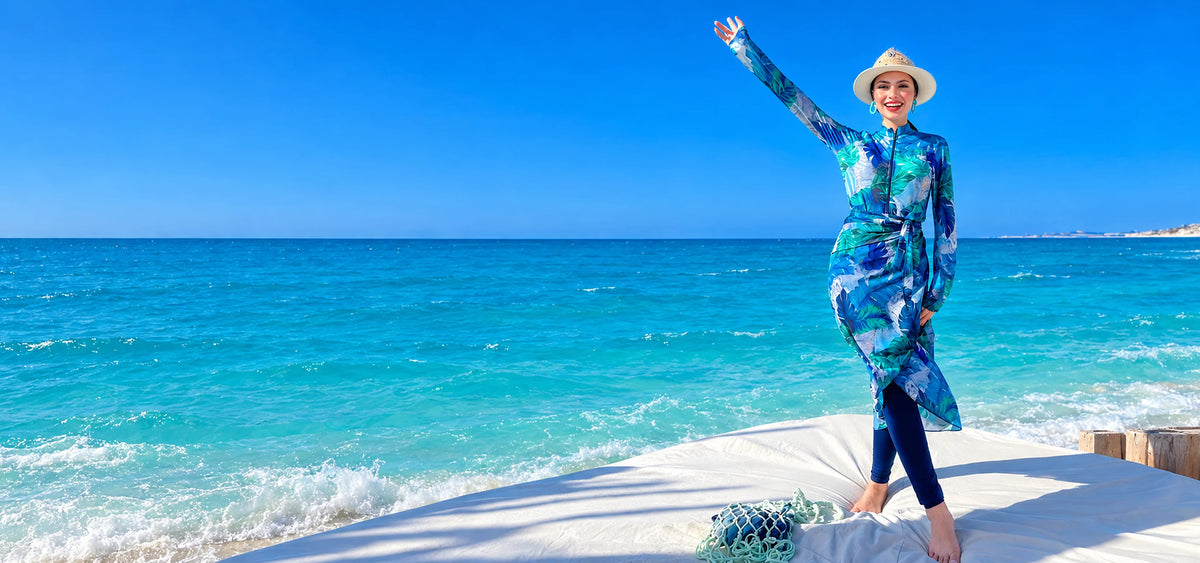 Smiling model in Tallah's blue botanical Burkini, matching wrap skirt and straw hat beside the sea.