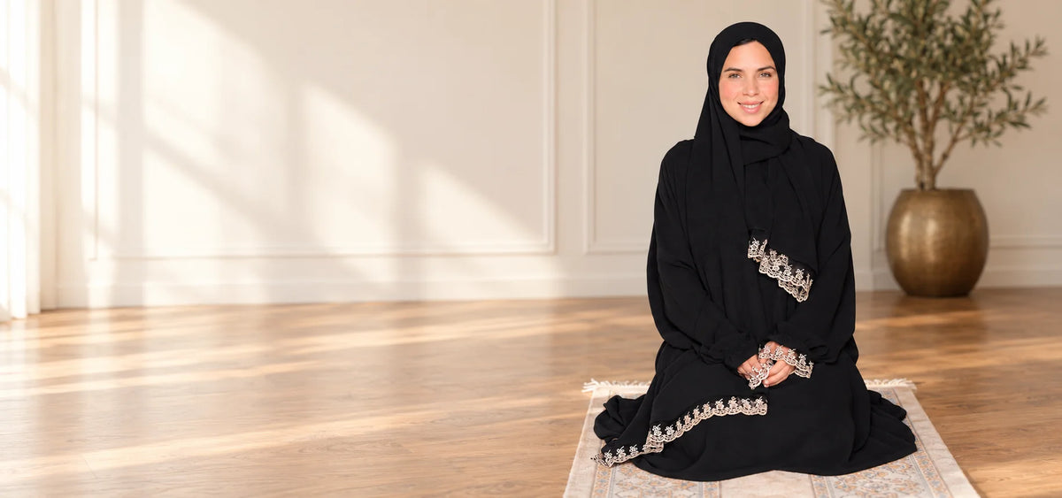 Woman wearing a black Isdal and hijab with ivory lace trim, seated on a prayer rug in a sunlit room.