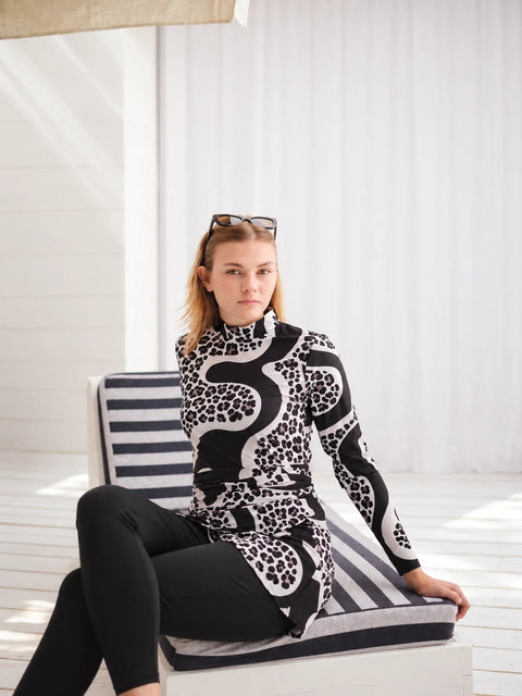 Seated model wearing a black and white patterned Burkini beside white curtains.