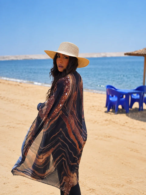 Model wearing a navy and rust marbled swim cover-up and a straw hat beside the sea.
