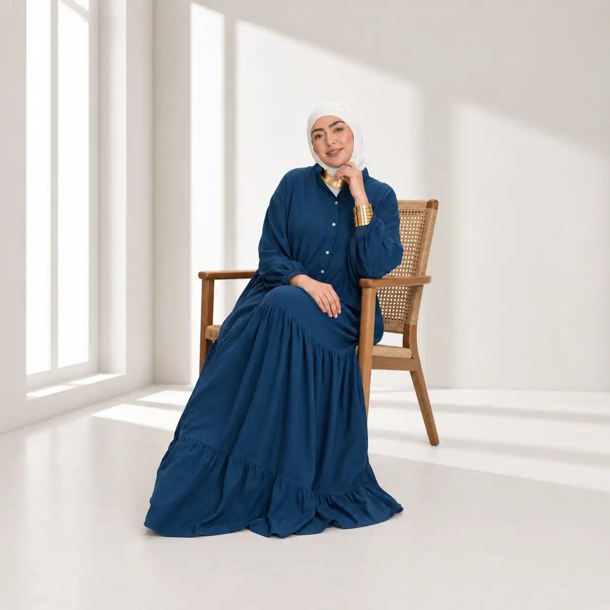 Model wearing a navy blue tiered maxi dress and white hijab, seated on a wooden cane chair in a sunlit room.