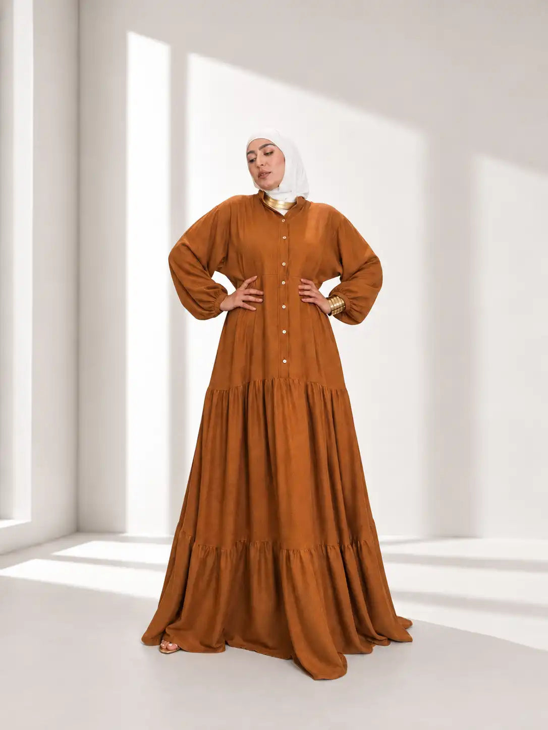 Model in a long caramel-brown tiered Tallah dress with hands on hips in a bright room