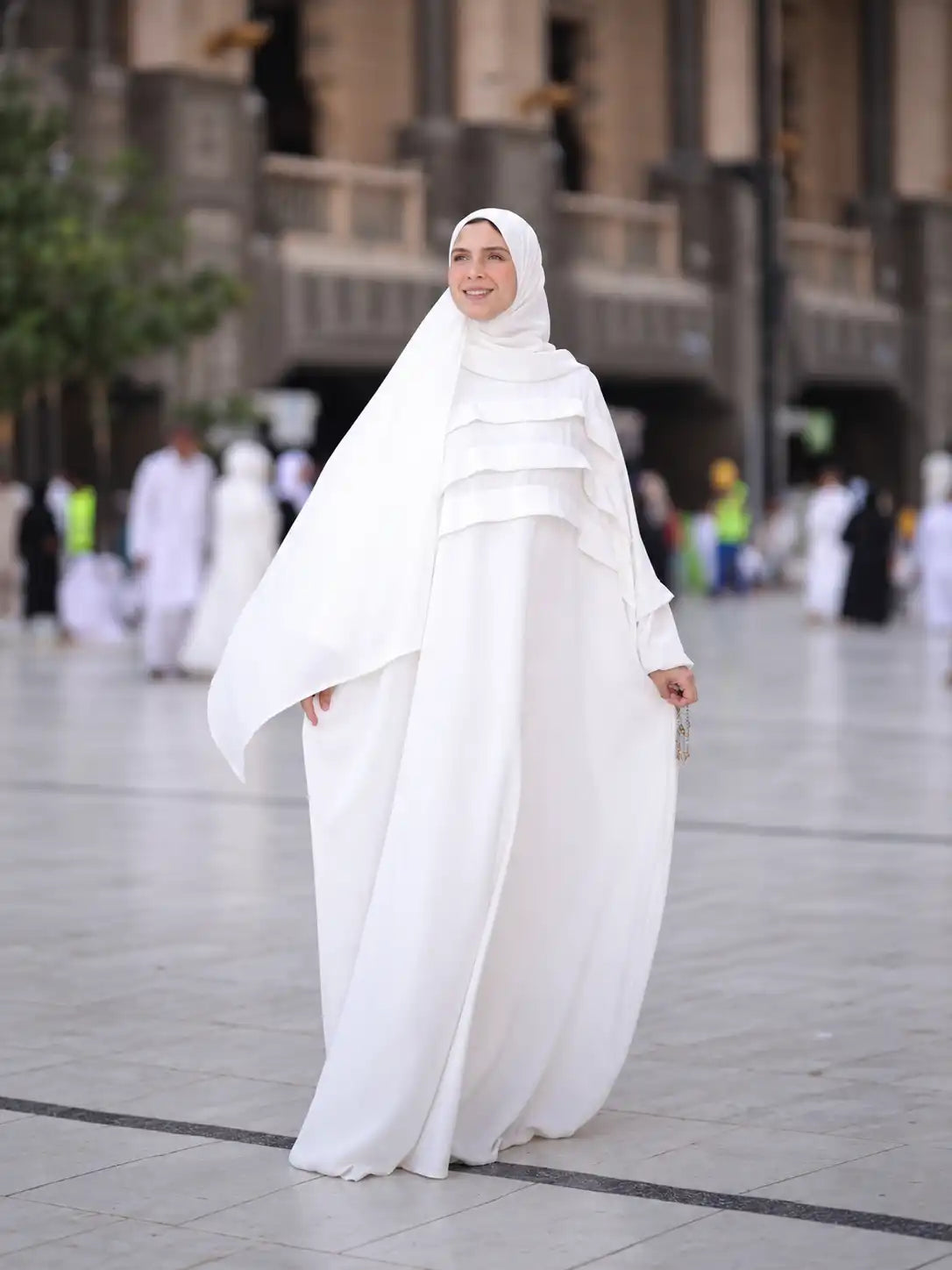 Model in a white flowing Tallah Hajj and Umrah prayer set with a headscarf, standing in a stone courtyard