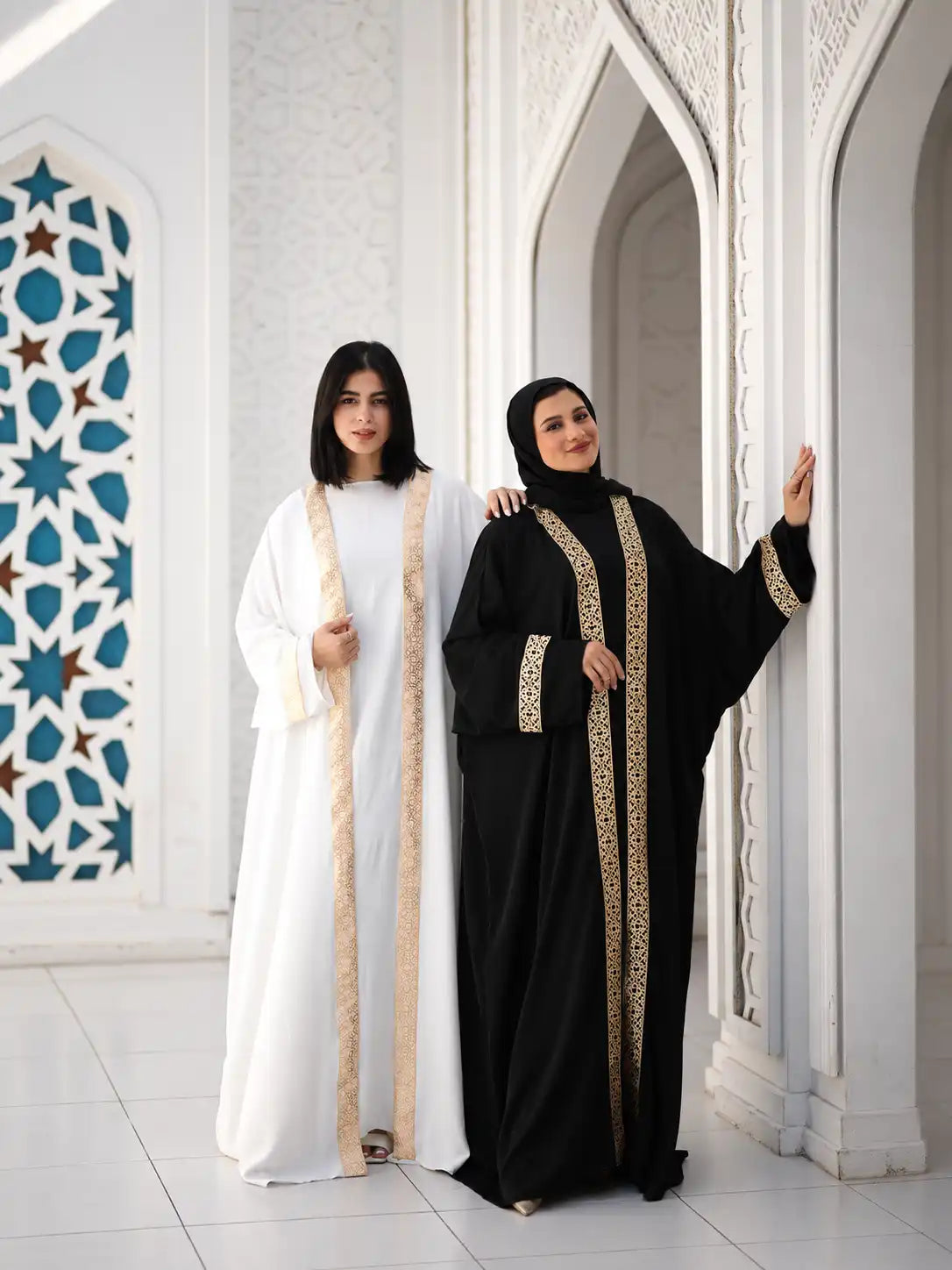 Two models in white and black Tallah kaftans with gold trim, standing in an arched doorway