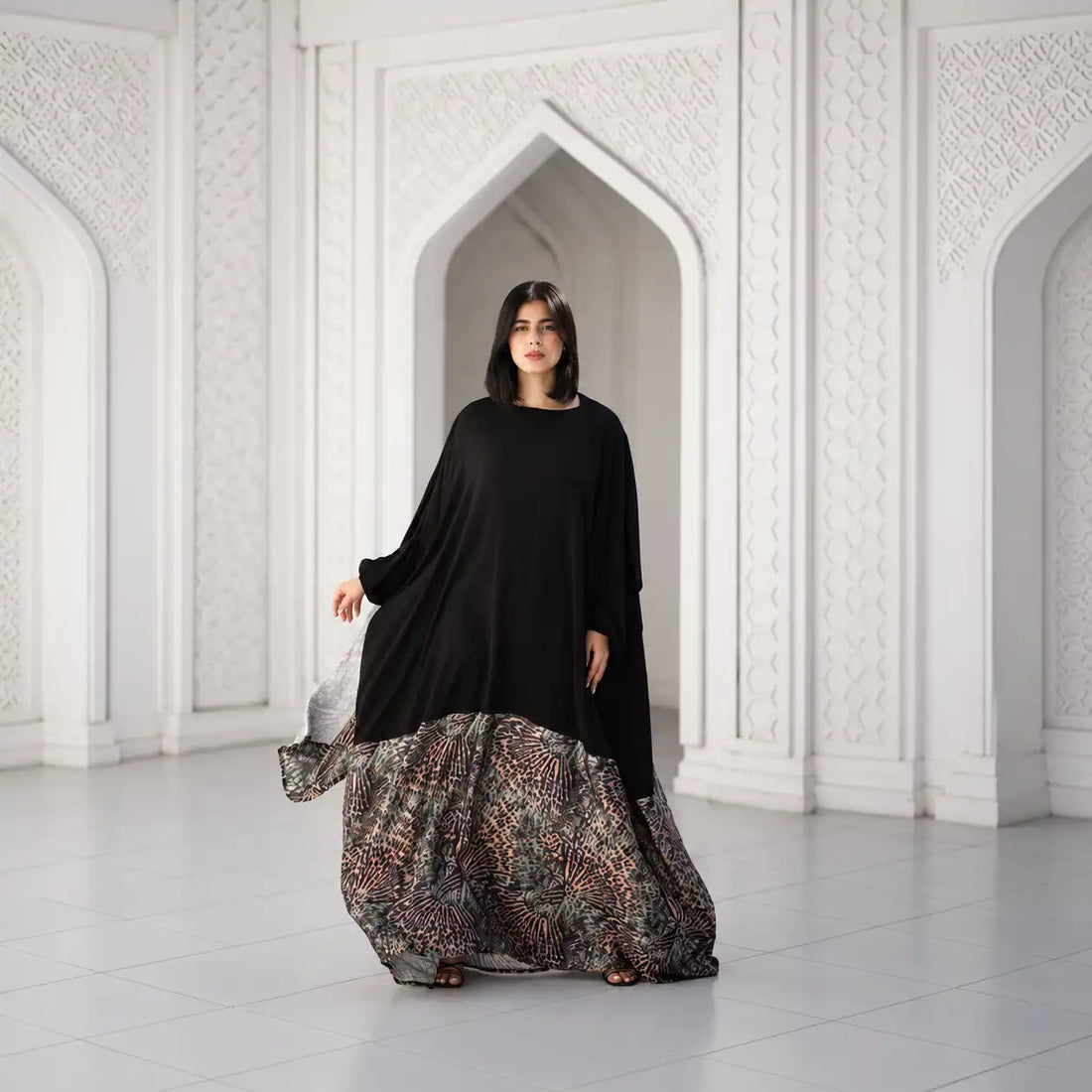 Model in a black Tallah Malhafa with a patterned skirt panel, standing in a white arched room