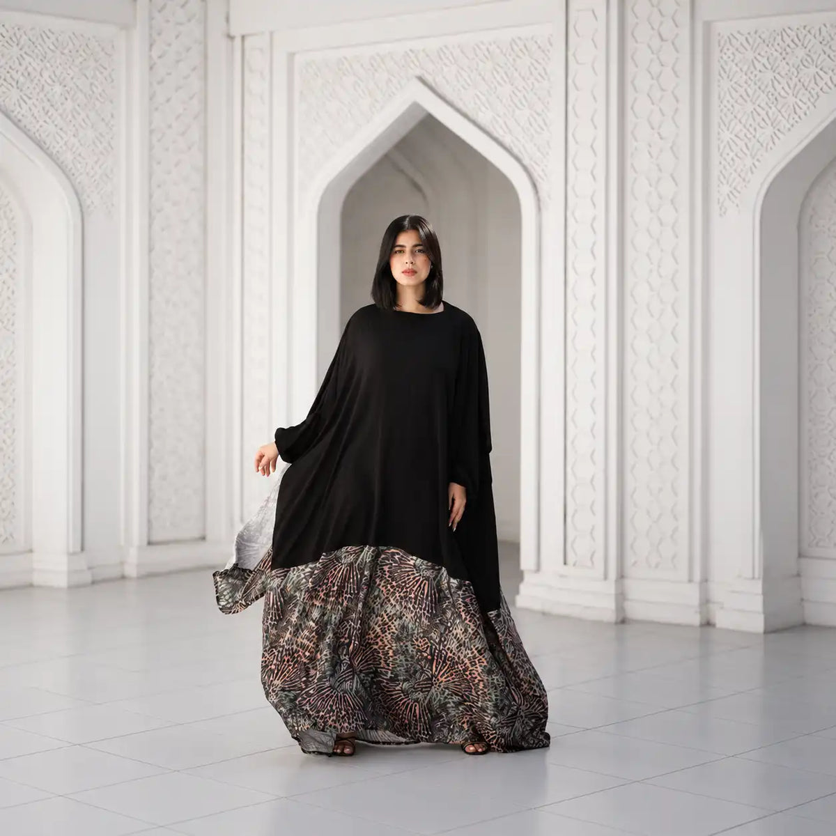 Model in a black Tallah Malhafa with a patterned skirt panel, standing in a white arched room