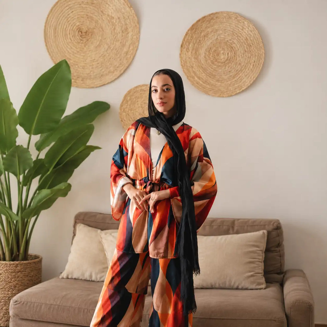 Model in a Tallah printed suit in orange, red and teal with a black hijab, standing in a living room