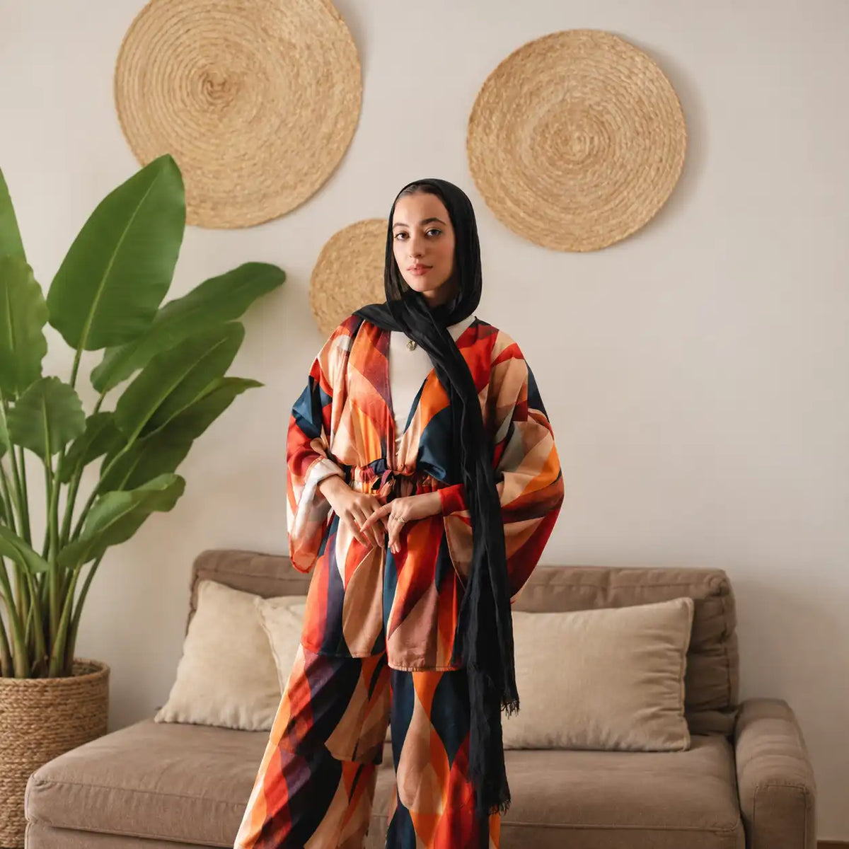 Model in a Tallah printed suit in orange, red and teal with a black hijab, standing in a living room