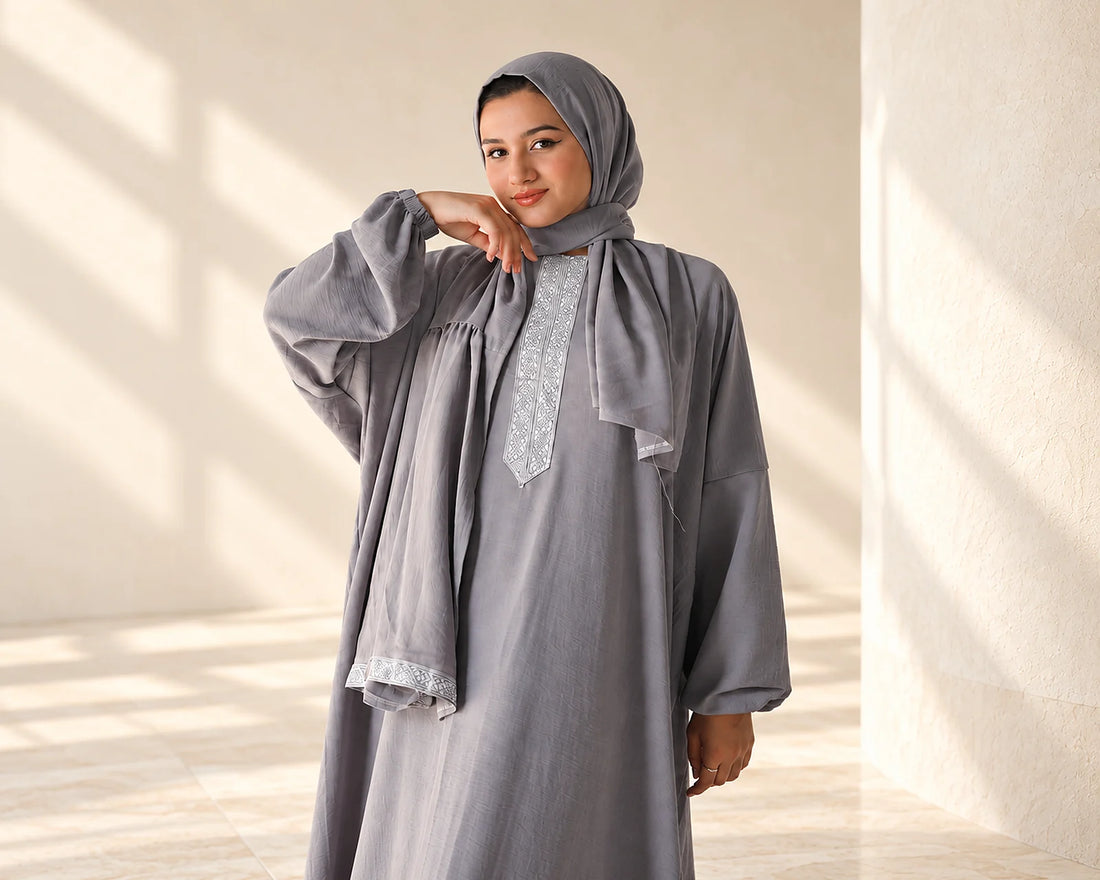 Model wearing a grey embroidered Isdal against a softly lit warm ivory stone backdrop.