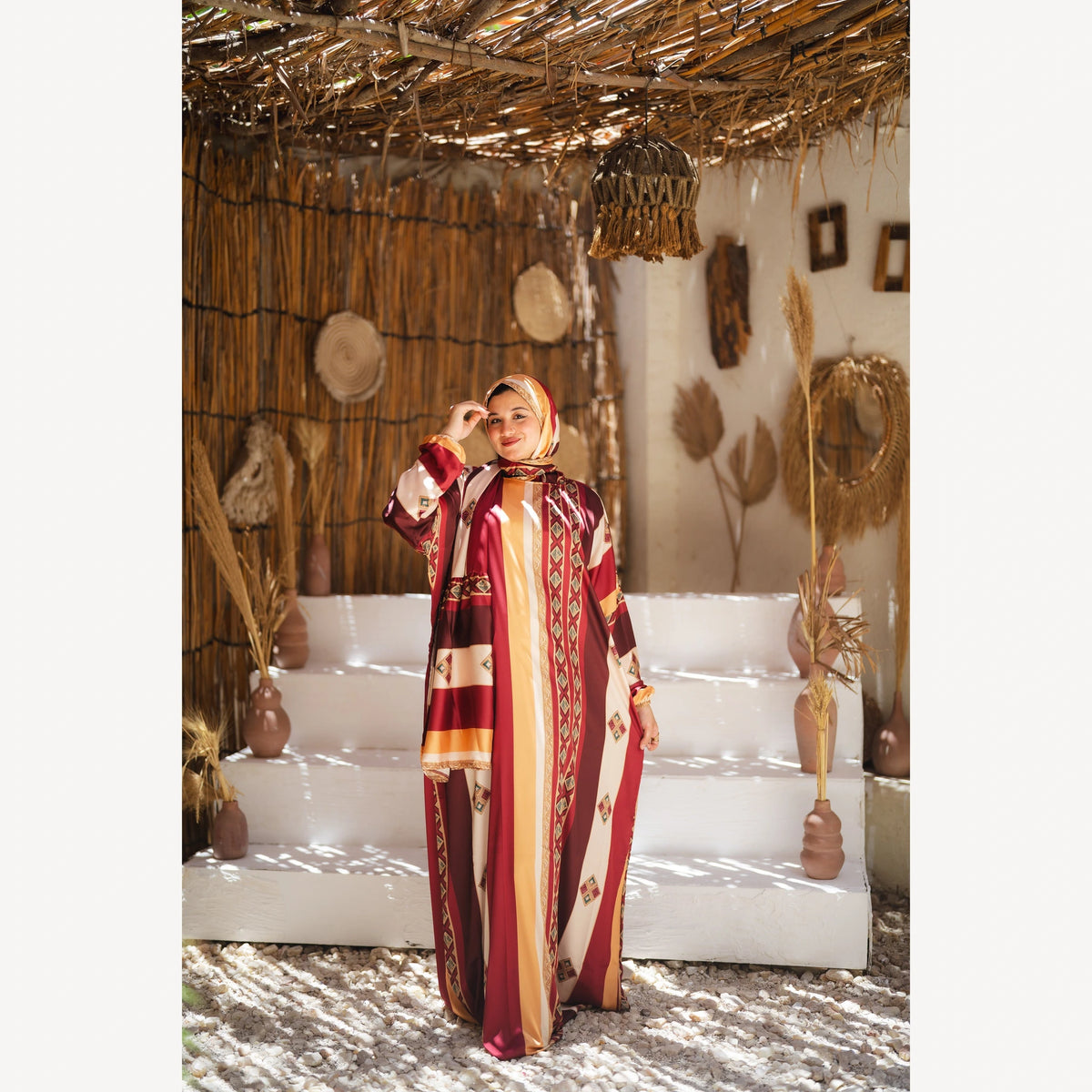 Tallah satin Isdal in burgundy mustard cream striped, full-length front view with one hand near forehead.