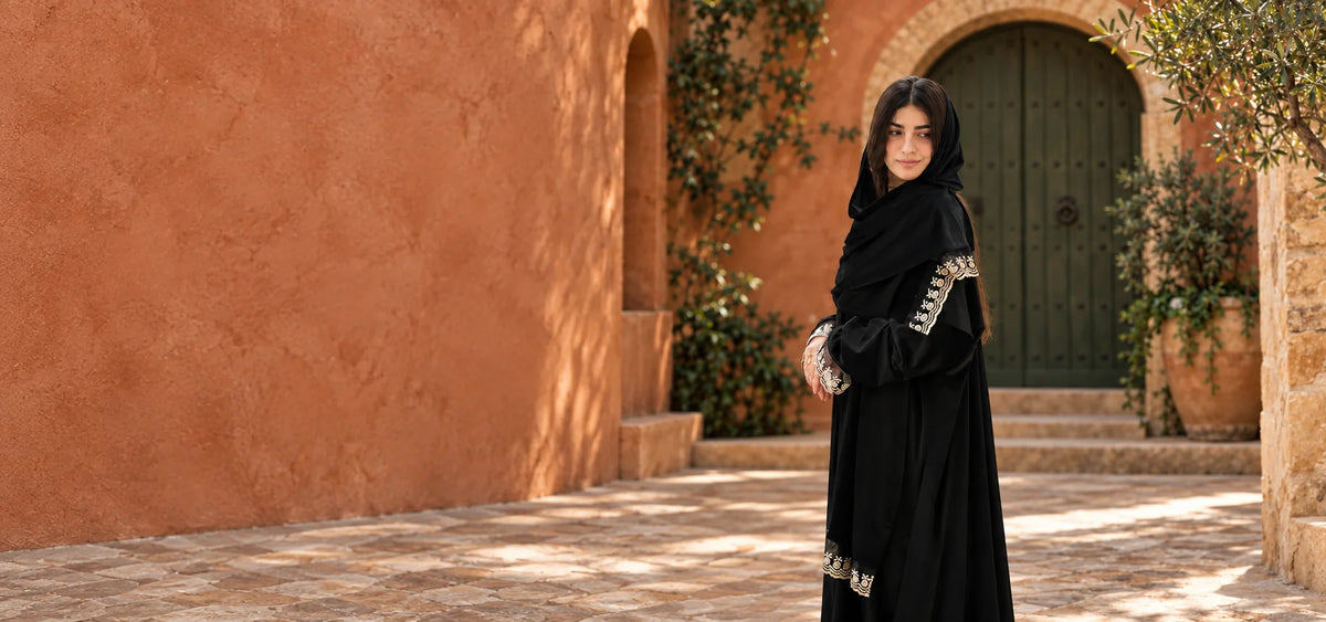 Woman wearing a black Isdal with ivory floral embroidery in a terracotta courtyard beside an olive-green door.